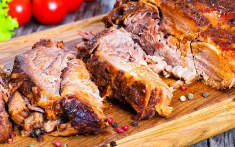 Une succulente épaule d'agneau rôtie, effilochée et servie sur une planche en bois, avec des tomates et de la laitue en arrière-plan.