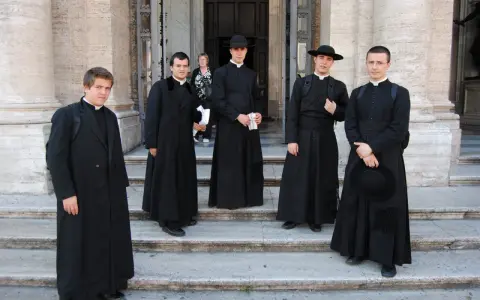 Cinque giovani seminaristi in abiti talari neri, alcuni con cappello, posano sulle scale di un edificio storico.