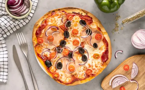 Gotowe ciasto na pizzę z szynką, oliwkami, pomidorkami i czerwoną cebulą. Obok sztućce i miseczka z cebulą.
