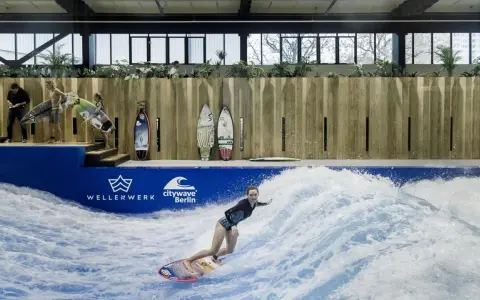 Frau surft auf einer künstlichen Welle in Berlin. Mehrere Surfbretter stehen bereit.