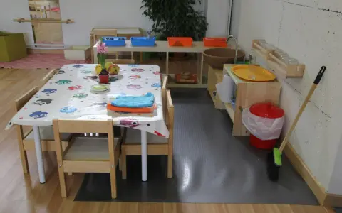 Mesa con mantel de búhos, toallas y frutas, lista para actividades en una casa de niños Montessori. Cerca, un lavabo y cubos para juegos.