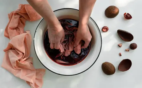 Mani che tingono tessuti in una bacinella con liquido scuro, circondata da bucce di avocado. Un processo naturale per come tingere i tessuti.