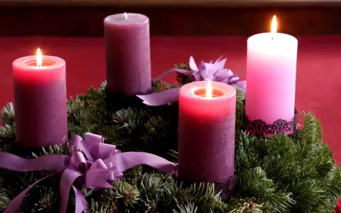 Drei brennende Kerzen auf einem Adventskranz aus Tannengrün mit lila Schleifen. Die dritte Kerze leuchtet hell.