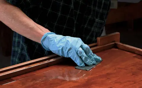 Mano enguantada lijando superficie de madera barnizada. El lijado es un paso clave para aplicar lac.