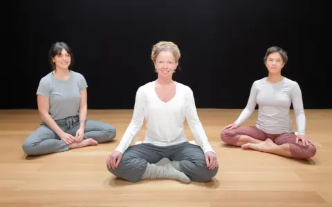 Tre kvinnor sitter i lotusställning på ett trägolv. De deltar i en klass i traumaanpassad yoga.