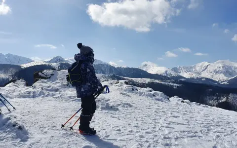 Chłopiec w zimowej kurtce i czapce z pomponem stoi na ośnieżonym szczycie, podziwiając wielki kopieniec gór. W rękach trzyma kijki trekkingowe.