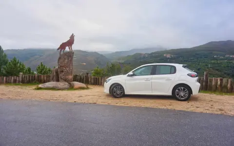Ein weißer Kleinwagen parkt neben einer Wolfsstatue auf einem Felsen mit Blick auf eine bergige Landschaft. Tolle Carjet Erfahrungen für diese Reise!