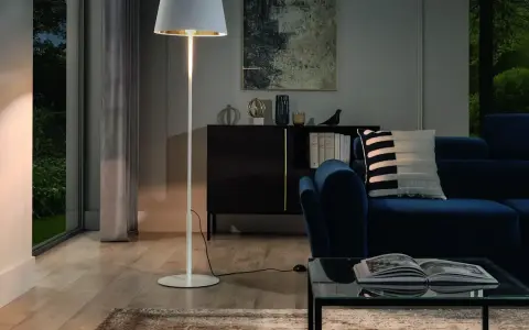 Biała, minimalistyczna lampa stojąca oświetla przytulny salon z granatową kanapą i szklanym stolikiem.