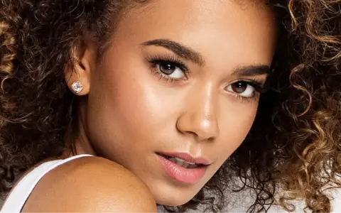 Un gros plan sur le visage d'une jeune femme aux cheveux bouclés, portant une boucle d'oreille étincelante. Ses **rehaucils avis** sont subtils, accentuant son regard.