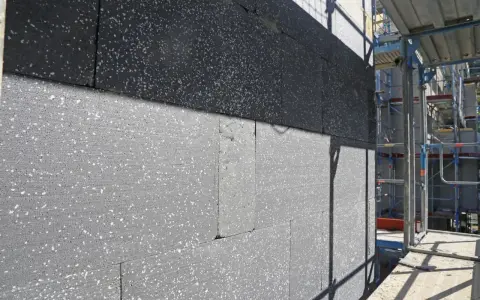 Gros plan sur des blocs de polystyrène gris, certains recouverts d'une couche noire, en cours d'installation pour une isolation par l'extérieur. Échafaudages visibles à droite.