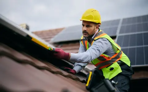 Instalator w żółtym kasku i kamizelce bezpieczeństwa montuje panele fotowoltaiczne na dachu. To dowód, że fotowoltaika jest bezpieczna, gdy montują ją profesjonaliści.