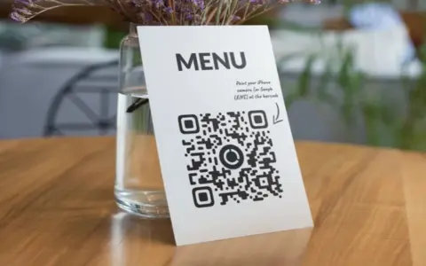 Un código QR de restaurante en una mesa de madera, junto a un jarrón con flores secas. Escanea para ver el menú.