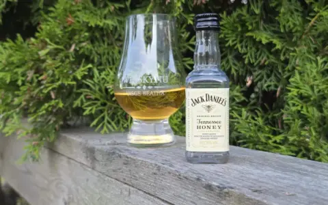 En liten flaska Jack Daniels Honey och ett whiskyglas står på en träbänk, omgivna av grönska.