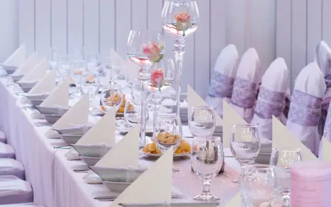 Vacker dukning med rosa rosor i glasvaser, vita servetter och silverfärgade skålar på en rosa bordsduk.