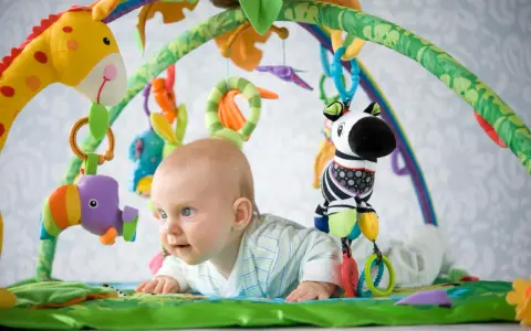 Niemowlę leży na kolorowej macie edukacyjnej, bawiąc się wiszącymi zabawkami: żyrafą, papugą i zebrą.