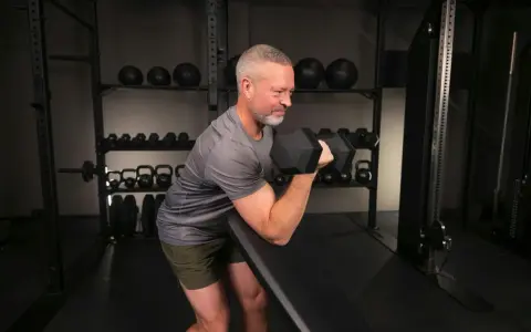 Man med grått hår och skägg utför preacher curls med en hantel i ett gym.