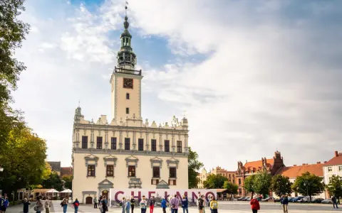 Rynek w Chełmnie z ratuszem i tablicą z napisem "Chełmno". Ludzie spacerują, podziwiając atrakcje miasta.