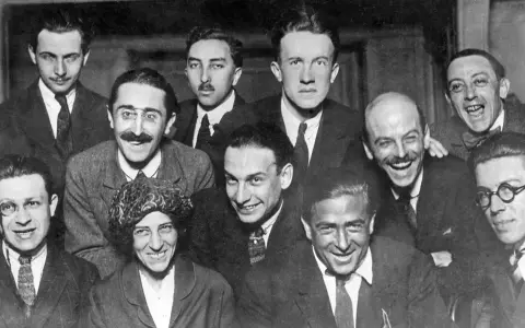 Un gruppo di uomini sorridenti, alcuni con baffi e occhiali, in una fotografia che evoca lo spirito irriverente del dadaismo.
