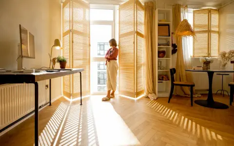 Una donna guarda fuori dalla finestra, mentre il sole crea ombre sul parquet che scricchiola. Un angolo studio e un tavolo da pranzo completano l'ambiente.