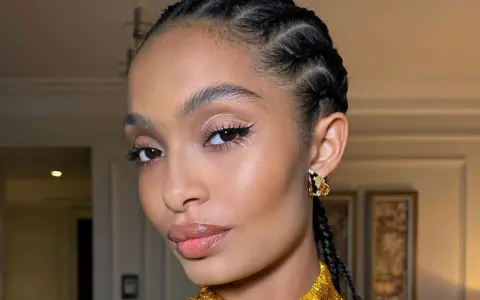 Primer plano de una mujer con trenzas, maquillaje de ojos con delineado y brillo, y labios brillantes.