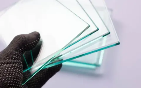 Mano enguantada sostiene varias láminas de vidrio laminar de seguridad, mostrando su transparencia y bordes verdosos.