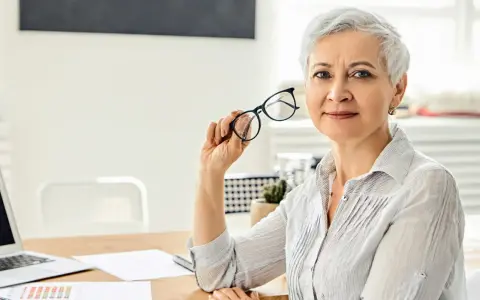 Mujer mayor con gafas, reflexionando sobre los tipos de jubilación. En su escritorio hay un portátil y documentos.