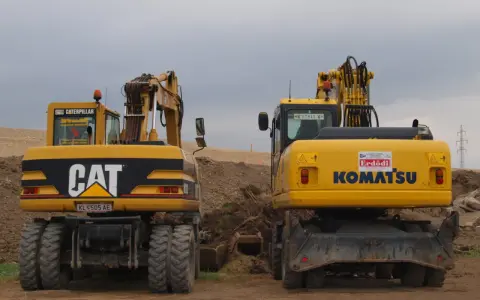 Dwie żółte koparki, CAT i Komatsu, stoją na placu budowy. Czy koparka ma dowód rejestracyjny? Na tablicy rejestracyjnej CAT widnieje KL 505 AE.