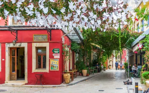 Ein rotes Gebäude mit weißen Tauben-Girlanden über einer Gasse in Chania, Kreta. Tische und Stühle laden zum Verweilen ein.