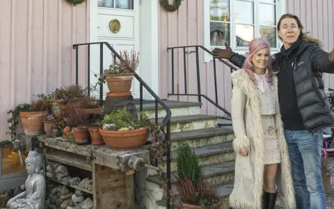 Melinda Jacobs och E-Type poserar glatt framför ett rosa hus med många krukor och en Buddha-staty.