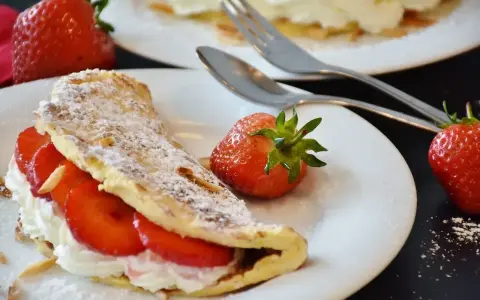 Słodki omlet z bitą śmietaną i truskawkami, posypany cukrem pudrem i płatkami migdałów. Idealny deser na słodko.
