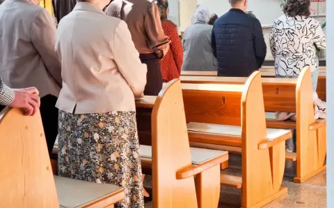 Ludzie stoją w kościele, czekając na dyspensę. Widać ławki i fragmenty ubiorów wiernych.