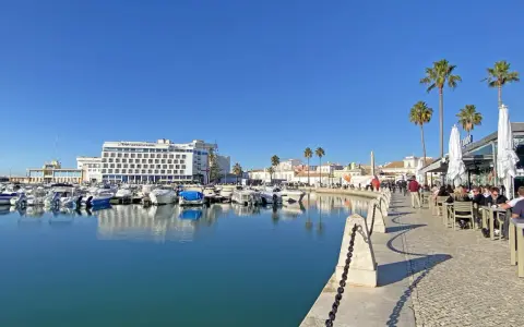 Boote im Hafen von Faro, Portugal, spiegeln sich im ruhigen Wasser. Palmen säumen die Promenade, wo Menschen in Cafés sitzen.