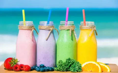 Cztery kolorowe smufi na plaży: różowe, fioletowe, zielone i żółte. Obok owoce: truskawki, maliny, borówki, pomarańcze i jarmuż.
