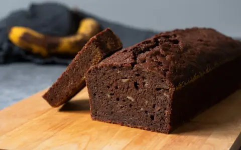 A slice of moist chocolate banana bread is propped against a whole loaf on a wooden cutting board.
