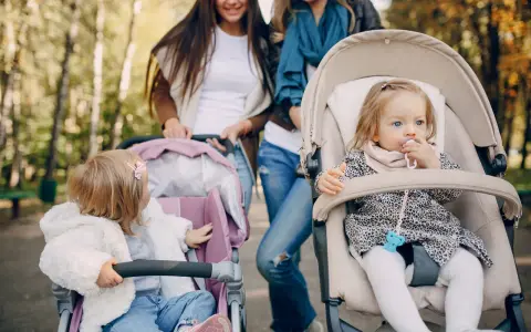 Dwie mamy spacerują z niemowlętami w wózkach. Jedno dziecko ma smoczek, drugie bawi się zabawką. Idealny spacer w parku.