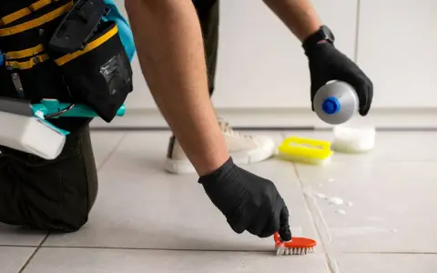 Persona con guantes negros usa un cepillo para limpiar juntas del suelo, aplicando producto de limpieza.