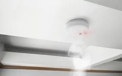 Detector de humo blanco en techo, emitiendo luz roja y humo. Cumpliendo la normativa de detectores de humo para seguridad.