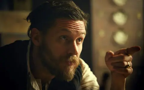 Mężczyzna z charakterystyczną fryzurą w stylu Peaky Blinders, z zarostem i pierścieniami na palcach, wskazuje palcem.