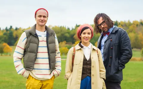 Tre personer i höstkläder står i en park. Mannen till vänster har en röd mössa och en randig tröja. Kvinnan i mitten bär en beige kappa och en rosa basker. Mannen till höger har glasögon och en fluga. De ser ut att vara med i "Geografens testamente".