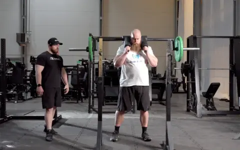 Man med skägg förbereder sig för knäböj med en safety squat bar. En tränare står bredvid.