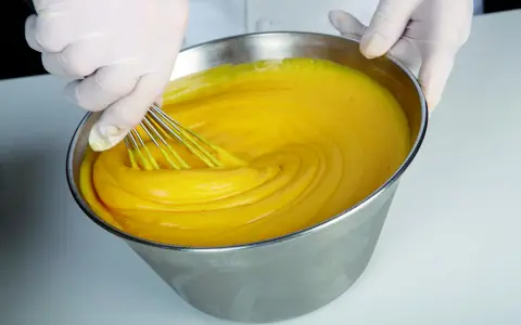 Mani guantate montano una crema pasticcera perfetta, come insegna Iginio Massari. La consistenza è vellutata e il colore giallo intenso.
