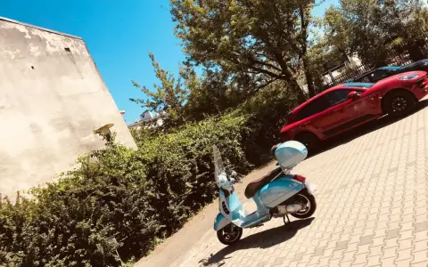 Błękitny skuter czeka na przejażdżkę. W tle czerwony samochód i zielone drzewa. Idealne miejsce na parking moto.