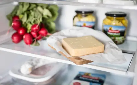 Ein Stück Käse liegt auf einem Holzbrett, daneben Radieschen und Gläser mit eingelegtem Gemüse. Verschiedene Käse sorten im Kühlschrank.