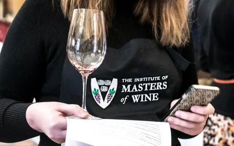 Eine Person mit einer Schürze des "Institute of Masters of Wine" hält ein Weinglas und ein Smartphone.