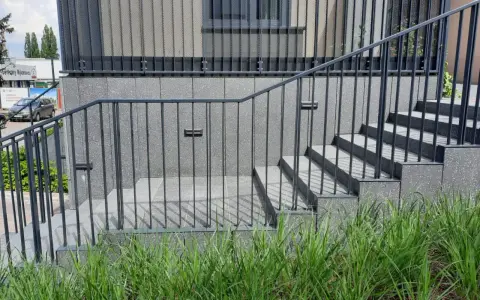 Nowoczesne schody zewnętrzne z metalową balustradą prowadzą do budynku. Wokół bujna zieleń.