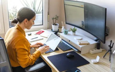 Mujer trabajando en un escritorio ordenado, con portátil, monitor curvo y plantas. Un ejemplo de como organizar tu escritorio para ser productivo.