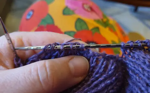 Närbild på en hand som stickar med lila garn. Nålen håller flera maskor, redo för nästa varv i blocka stickning.