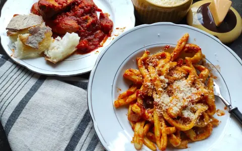 Pyszne maccheroni al ferretto w sosie pomidorowym, posypane parmezanem, obok talerz mięsa w sosie i chleba.