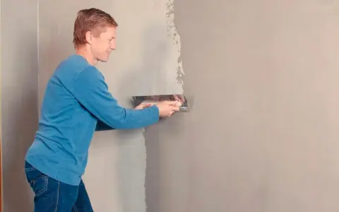 Man med blå tröja och jeans använder en bred spackel för att rolla spackel på en vägg.