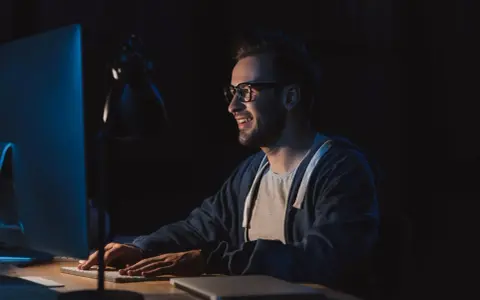 Uśmiechnięty mężczyzna w okularach pracuje nocą przy komputerze, przygotowując się do studiów podyplomowych tester oprogramowania.